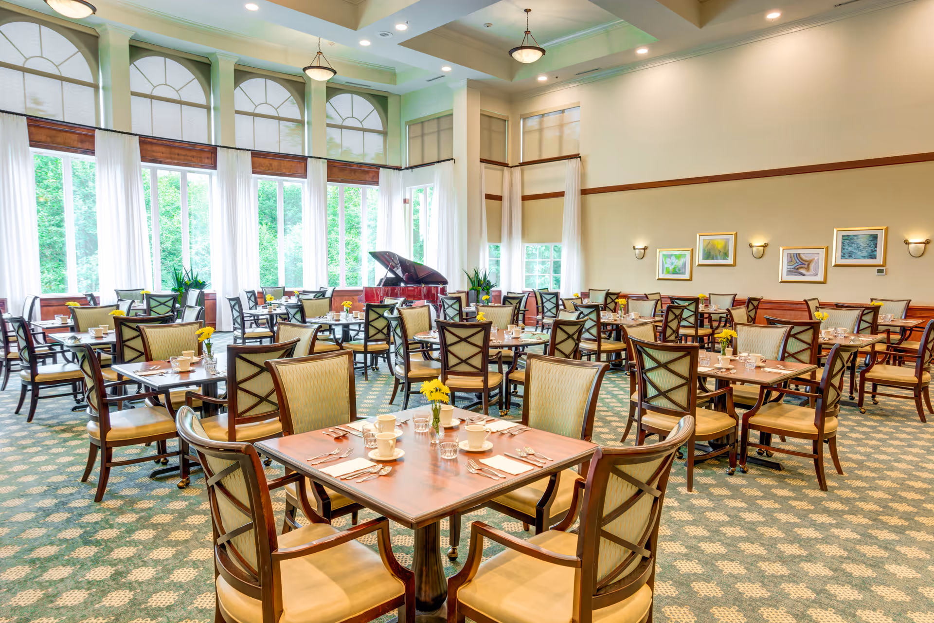 A bright and spacious dining room with multiple wooden tables and cushioned chairs arranged neatly. Each table is set with cups, glasses, and silverware, and small yellow flower centerpieces. Large windows with white curtains allow natural light to fill the room. A grand piano is positioned near the windows, and framed artwork decorates the walls.