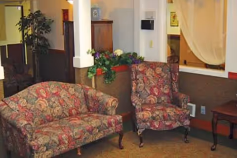 A cozy seating area in a memory care facility featuring a floral-patterned loveseat and armchair, a small wooden side table, a planter with green and purple plants, and a partially visible curtained window.