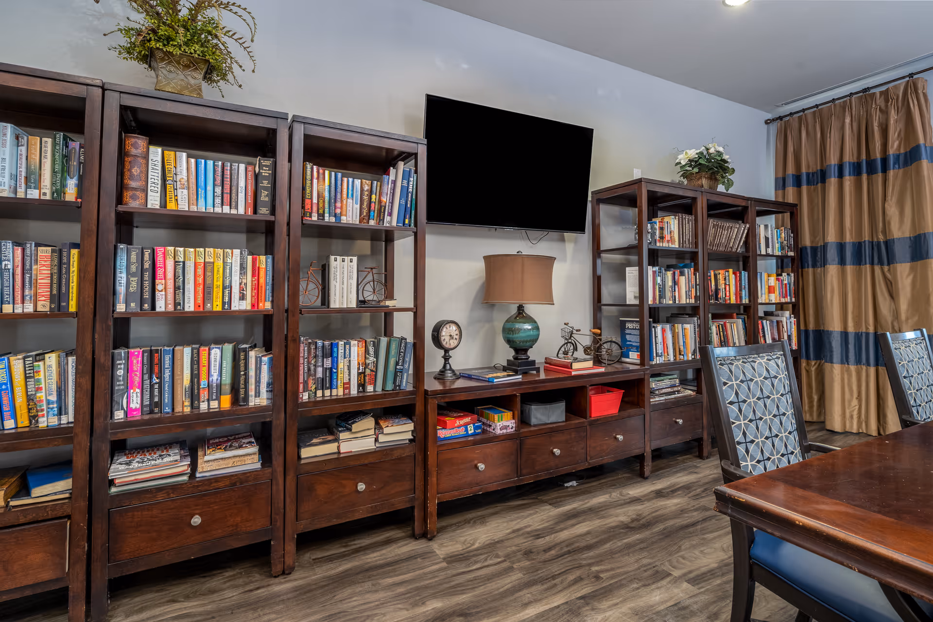 A cozy interior room featuring multiple dark wooden bookshelves filled with books and decorative items. A flat-screen TV is mounted on the wall above a wooden console table with a lamp, clock, and small bicycle decorations. There are patterned chairs and a wooden table in the foreground, with beige and blue striped curtains on the right side.