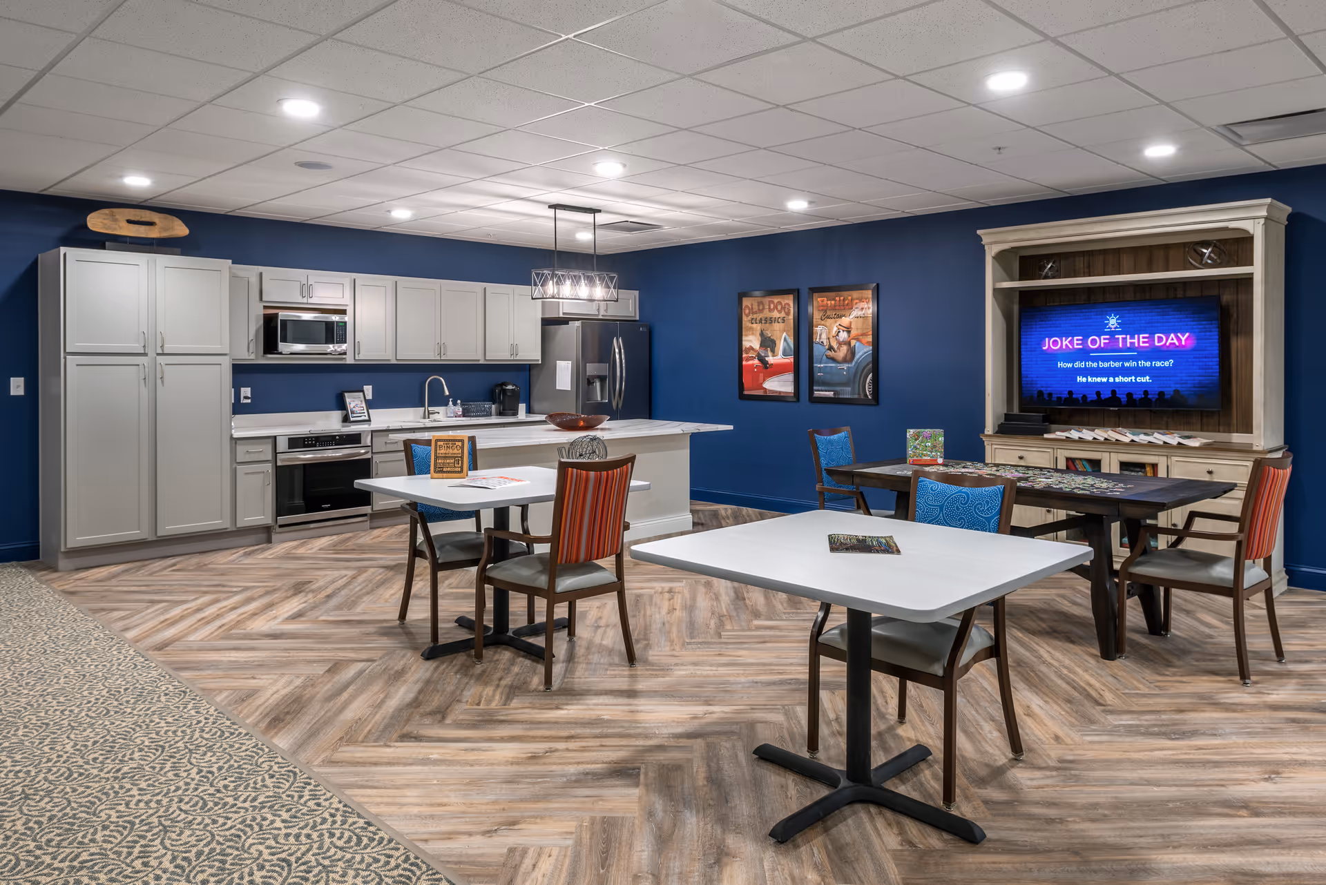 A common area with a kitchen and seating space. The kitchen has gray cabinets, a microwave, oven, and refrigerator. There are several tables with chairs around them, some with colorful cushions. A large TV on a wooden entertainment unit displays a 'Joke of the Day'. The walls are painted dark blue, and the floor has a wood pattern with a carpeted section on the left.