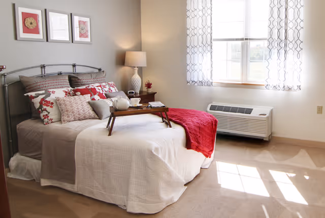 Sunlit bedroom with a made bed topped with decorative pillows and a red throw, a bedside table and lamp, and a window with patterned curtains.