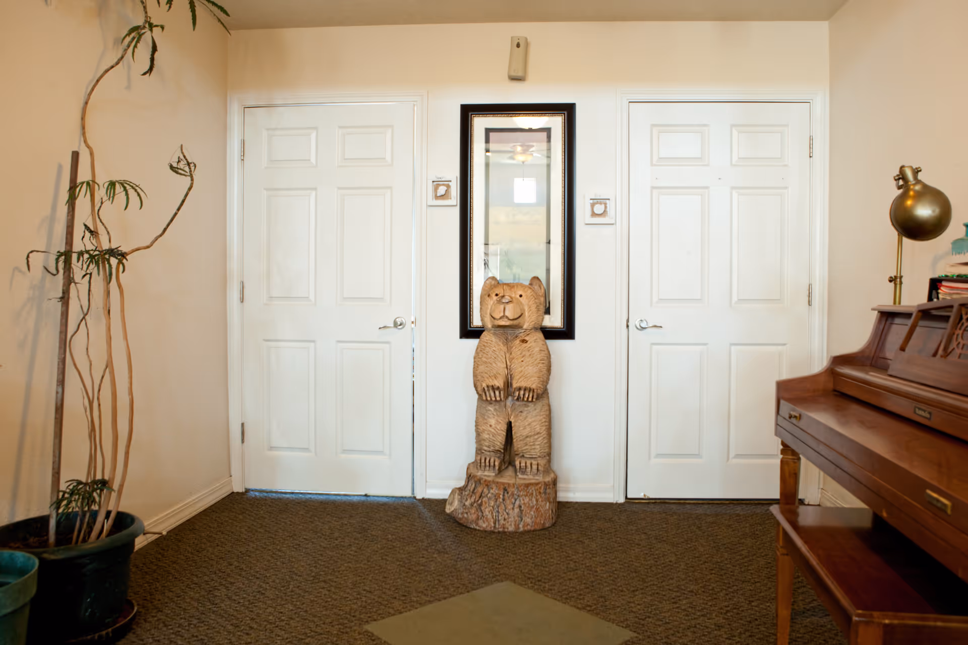 Entryway with two white doors, a carved wooden bear statue beneath a mirror, a piano on the right and potted plants on the left.