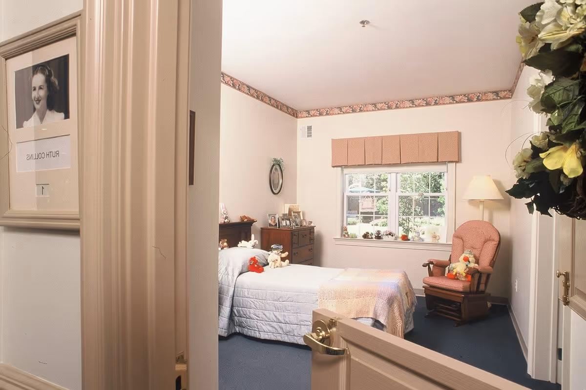 View through a partially open door into a cozy bedroom with a single bed covered with a light blue quilt and a pastel-colored blanket. The bed has several stuffed animals on it. There is a wooden dresser with framed photos and decorations, a window with a beige valance letting in natural light, and a pink cushioned rocking chair with stuffed animals on it. The room has floral wallpaper border near the ceiling and a lamp beside the chair. A framed black and white portrait and a nameplate reading 'Ruth Collins' are visible on the door.