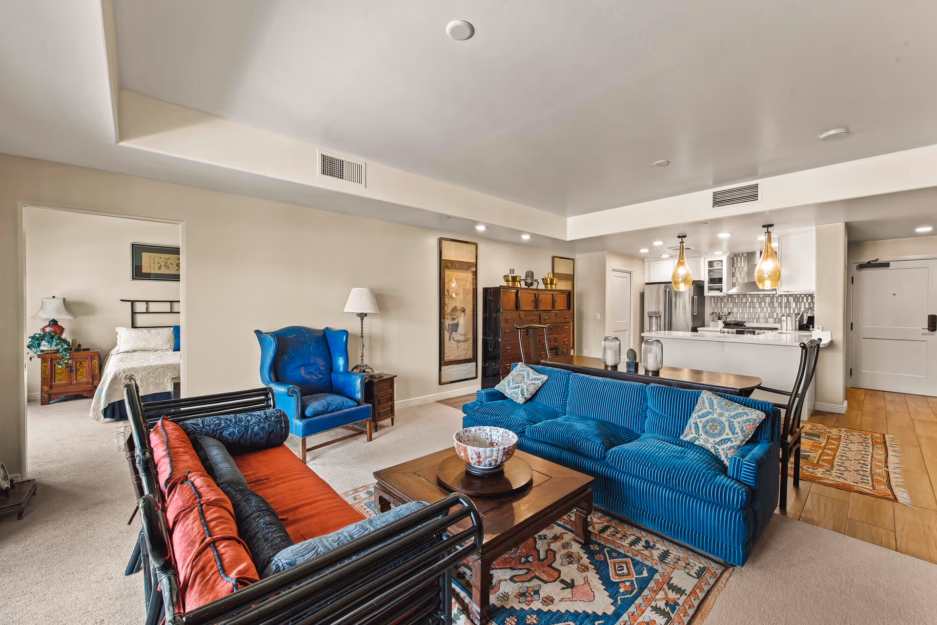Open-concept living room with blue sofas, patterned rug and wooden coffee table, with a view into the kitchen and a bedroom doorway.
