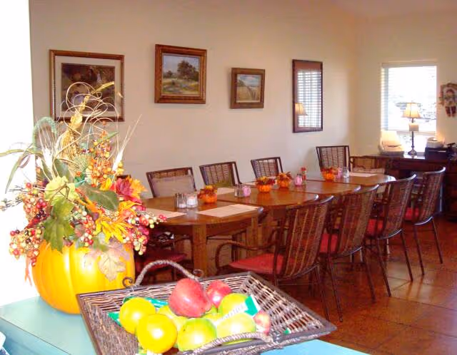 A dining room with a long wooden table surrounded by chairs with red cushions. The table is decorated with small pumpkin centerpieces and place settings. On the left side, there is a large pumpkin vase filled with autumn-themed flowers and berries. In the foreground, a basket contains various fruits including apples and lemons. The walls have framed landscape paintings, and there is a window with blinds letting in natural light.