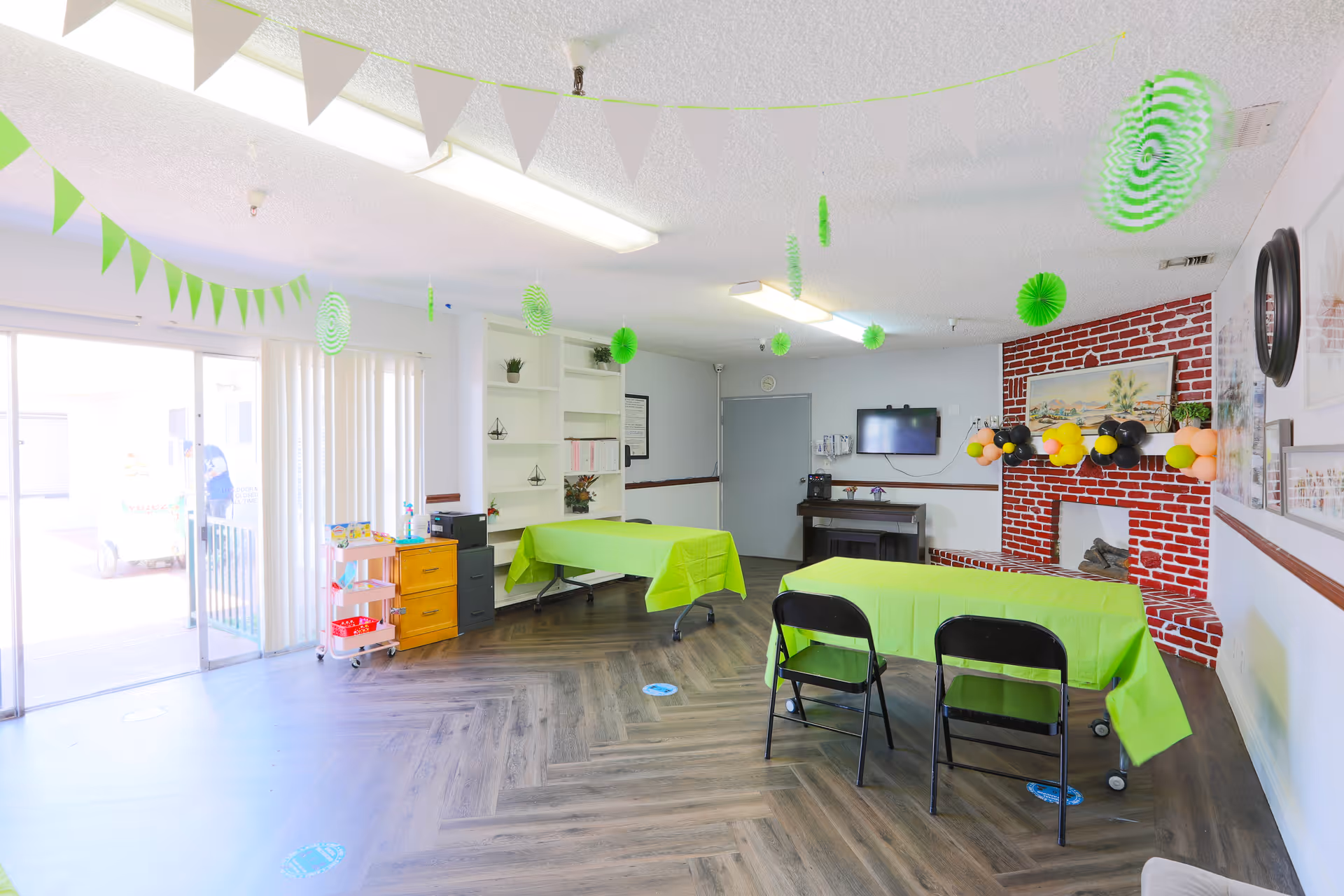 Bright multipurpose common room with tables covered in green tablecloths, folding chairs, a wall-mounted TV, and festive green decorations.