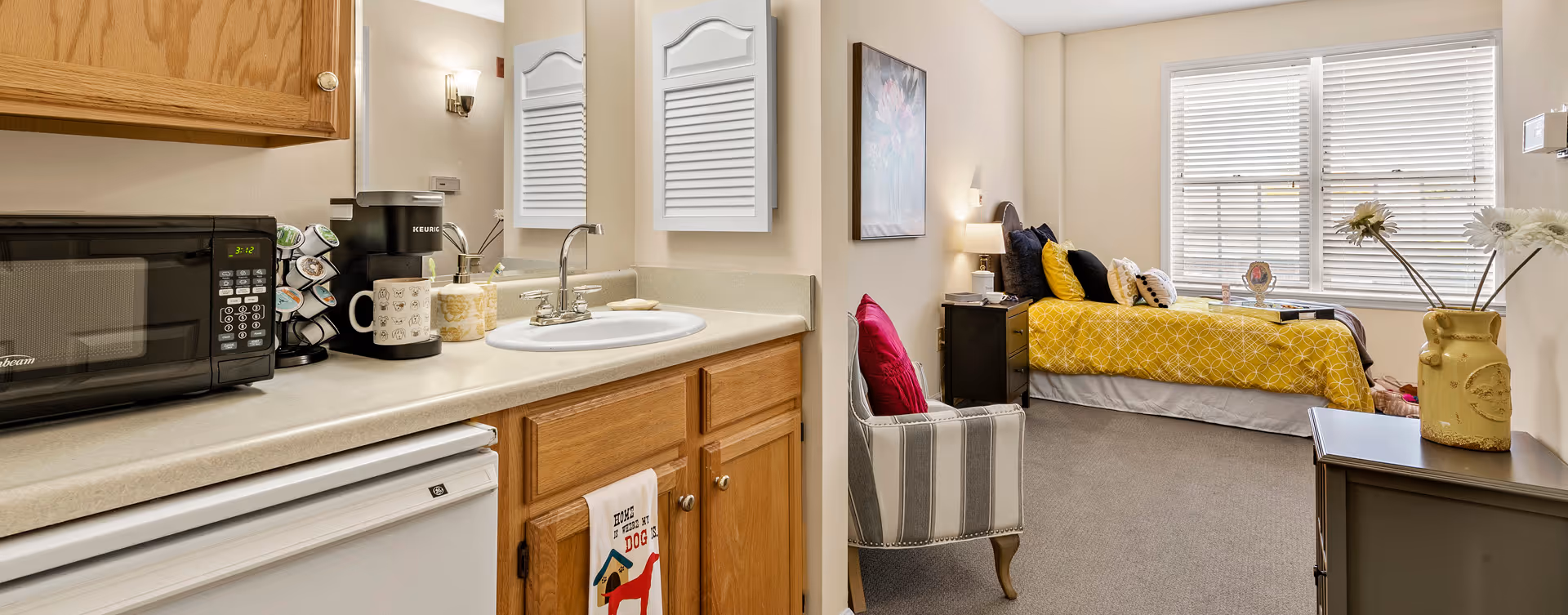 A cozy senior living facility room featuring a small kitchenette with a microwave, coffee maker, and sink on the left side. The room extends into a bedroom area with a bed covered in a yellow patterned comforter, multiple pillows, a nightstand with a lamp, a striped armchair with a red pillow, and a window with white blinds letting in natural light. A vase with white flowers is placed on a small table near the window.