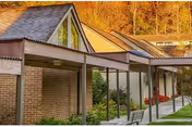 Exterior view of a senior living facility named Broadmead, showing a covered walkway with a sloped roof, brick walls, large windows, and a bench along the path. Trees with autumn-colored leaves are visible in the background.