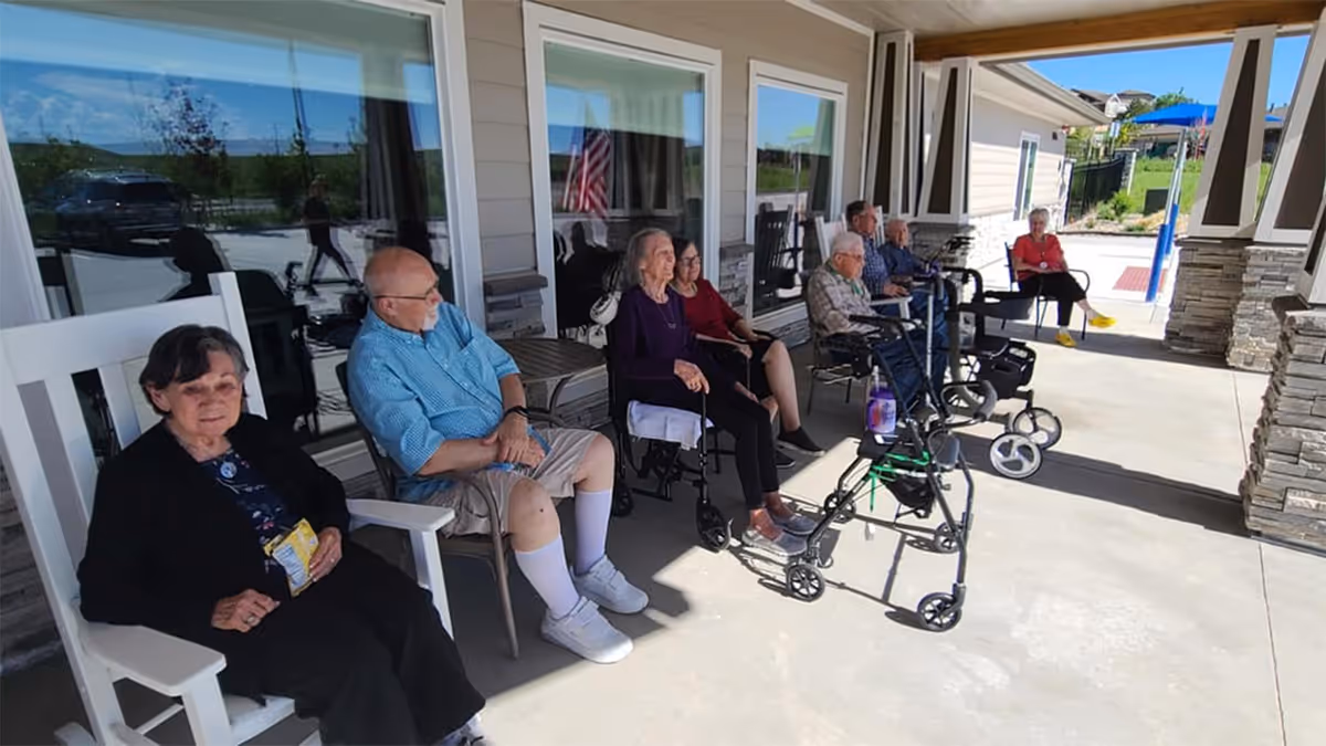 Several elderly residents seated on a covered porch outside a senior living facility, some using walkers and rocking chairs.