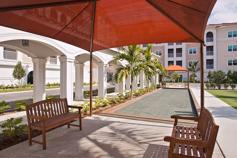 Outdoor seating area with wooden benches under red shade canopies next to a bocce ball court, surrounded by palm trees and landscaping, with a multi-story building in the background.