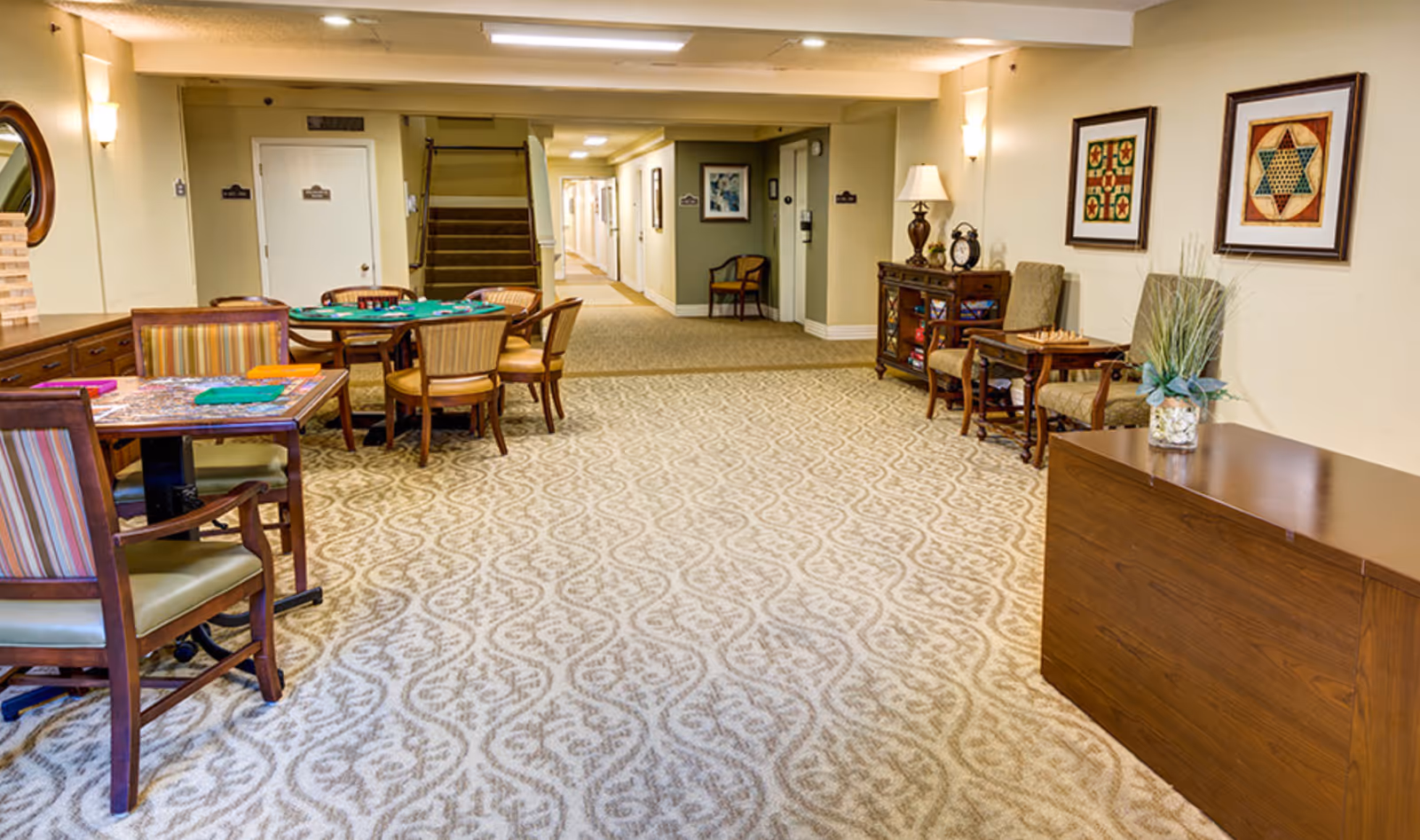 A well-lit common area in a senior living facility featuring patterned carpet flooring, several wooden tables and chairs arranged for games and social activities, framed artwork on the walls, a wooden cabinet with a lamp and clock, and a hallway leading to other rooms and a staircase.