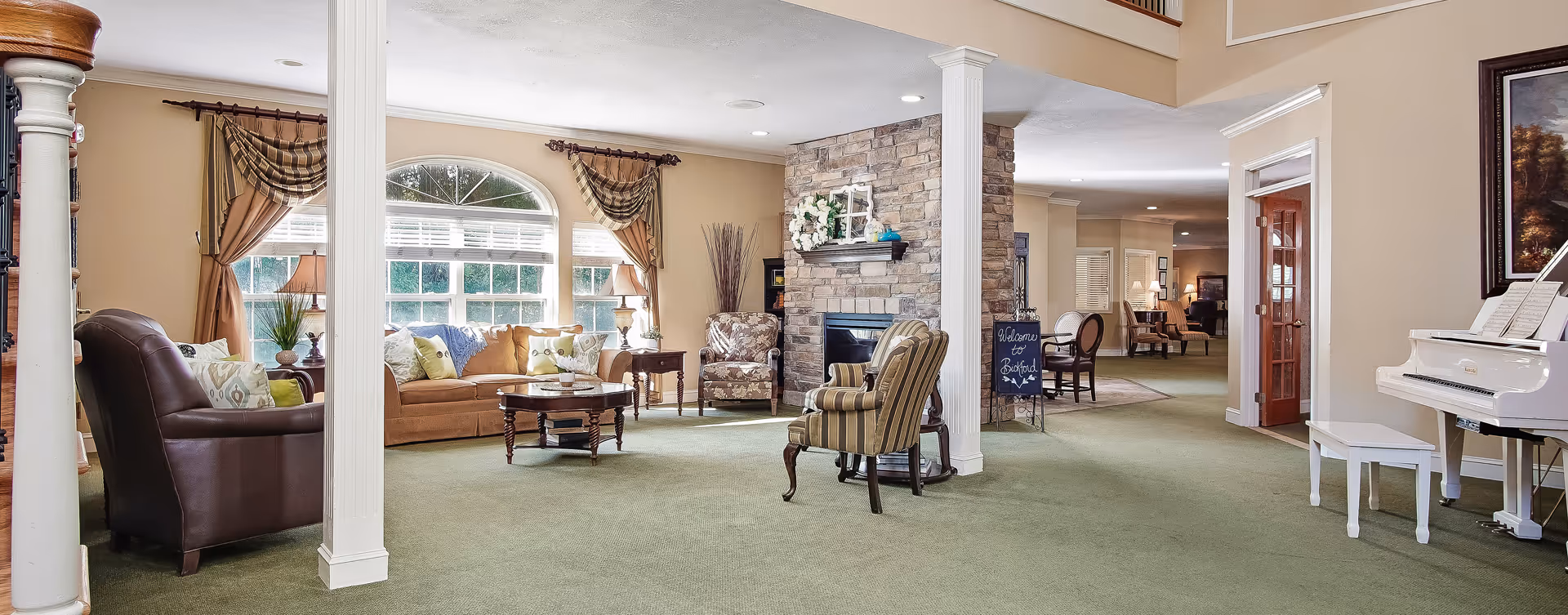A spacious and well-lit living room area in a senior living facility featuring a variety of comfortable seating including a brown leather armchair, a beige sofa with decorative pillows, and patterned armchairs. The room has large windows with draped curtains, a stone fireplace with a wreath and decorative items on the mantel, a white piano with a bench, and a small chalkboard sign that reads 'Welcome to Bickford'. The walls are painted beige and the floor is covered with green carpet.