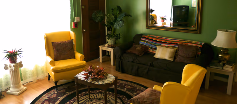 A cozy living room with two yellow armchairs and a dark sofa adorned with multiple cushions and a colorful crocheted throw. A small wooden coffee table with a floral centerpiece sits on a patterned round rug. The room has green walls, a large mirror above the sofa, a table lamp on a side table, and a potted plant near a wooden door. Light filters in through a window with sheer curtains.