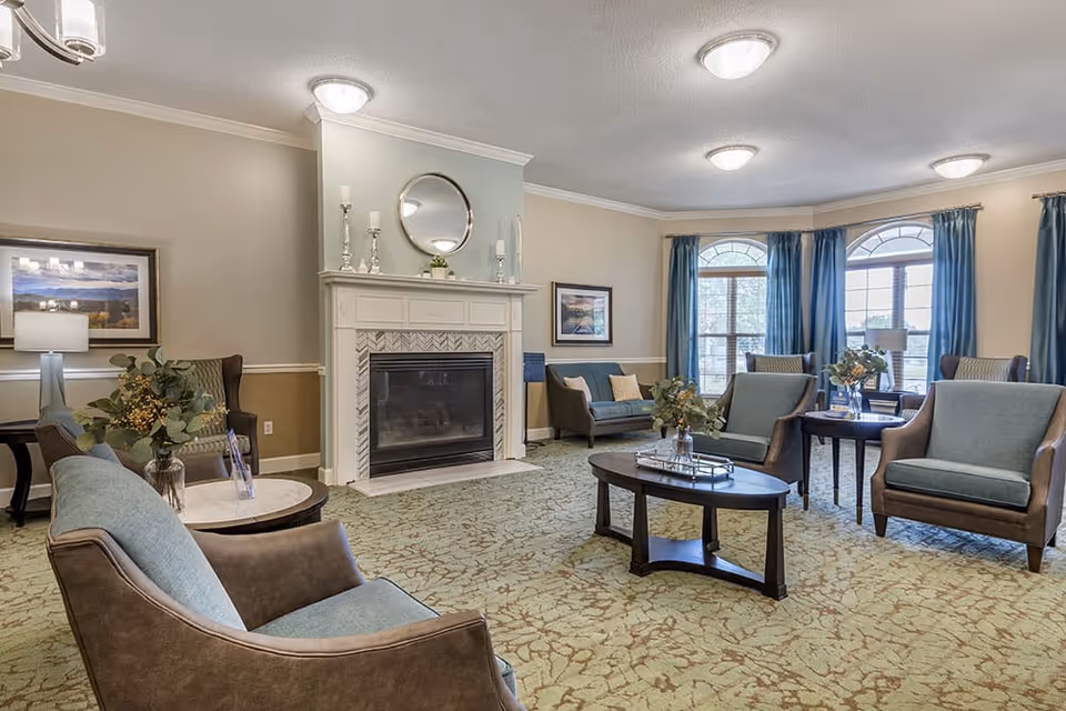 A spacious living room with multiple armchairs and sofas arranged around a central coffee table. The room features a fireplace with decorative candlesticks and a round mirror above it. Large windows with blue curtains allow natural light to fill the space, and framed artwork hangs on the walls. The carpet has a subtle pattern, and there are side tables with lamps and floral arrangements.