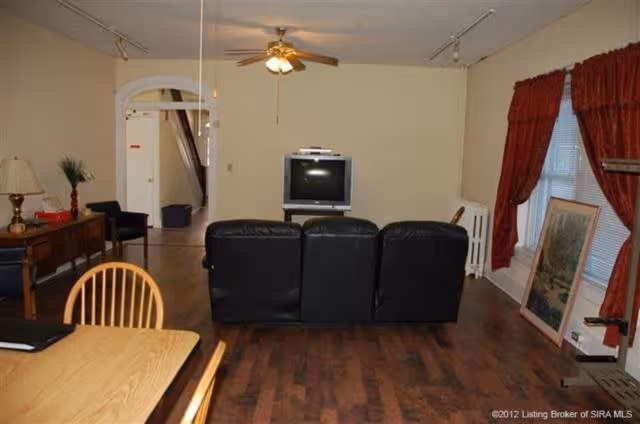 Interior view of a living room area with a black leather couch facing an old-style CRT television on a stand. The room has wooden flooring, a ceiling fan with lights, and beige walls. To the right, there is a window with red curtains and a framed picture leaning against the wall. On the left side, there is a wooden dining table with chairs and a side table with a lamp and decorative items. An open doorway leads to another room in the background.