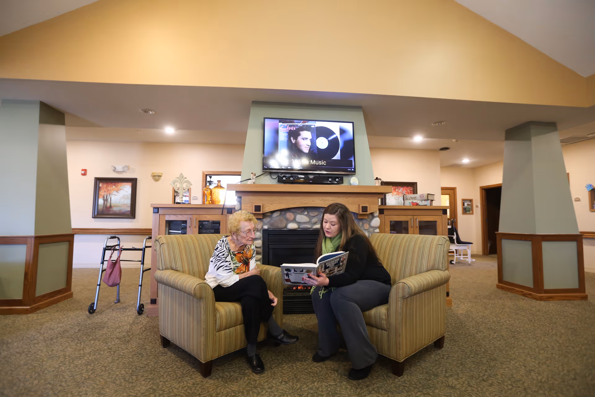 A staff member reads to an elderly woman seated on chairs in a cozy memory care living room with a fireplace and TV.