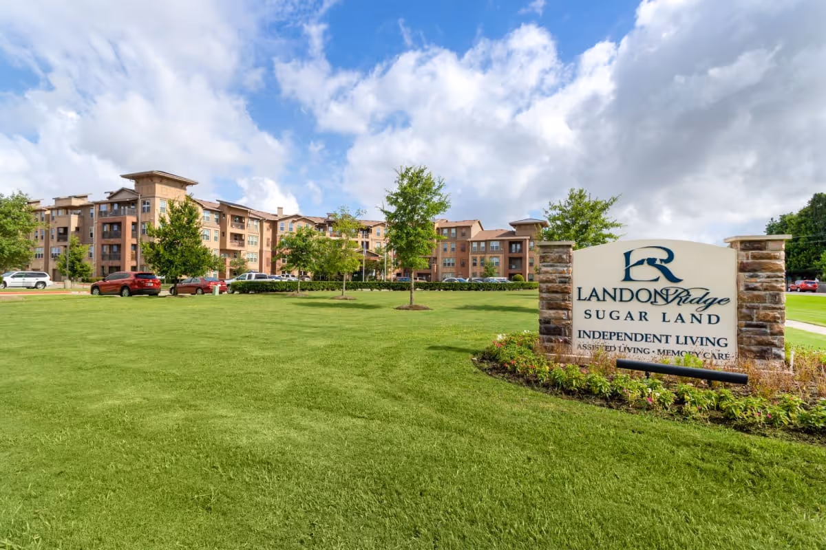 A large green lawn with a few young trees and a stone sign that reads 'Landon Ridge Sugar Land Independent Living Assisted Living Memory Care' in front of a multi-story residential building under a partly cloudy sky.