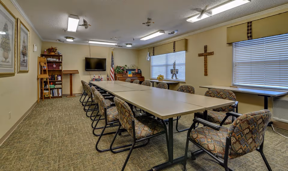 A meeting or activity room with a long rectangular table surrounded by patterned cushioned chairs. The room has beige walls, carpeted floor, two windows with blinds and valances, a wooden cross on the wall, an American flag, a TV mounted on the wall, a bookshelf with books and decorations, and a podium.