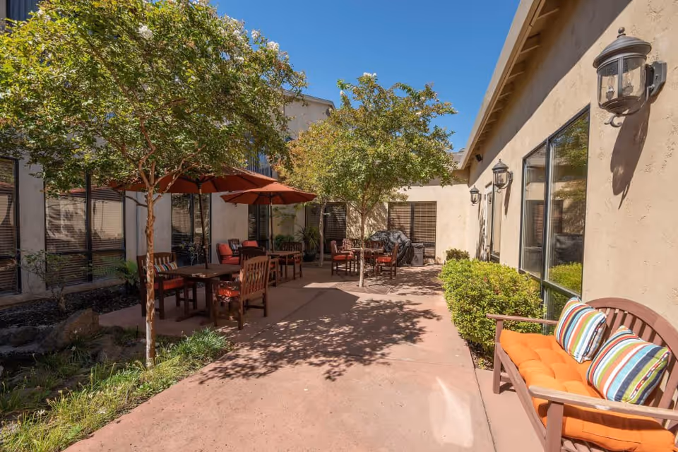Outdoor patio area at Winding Commons Senior Living with several wooden tables and chairs, some shaded by red umbrellas. There are small trees providing additional shade, a wooden bench with orange cushions and striped pillows, and wall-mounted lantern-style lights on the building exterior. The sky is clear and blue.