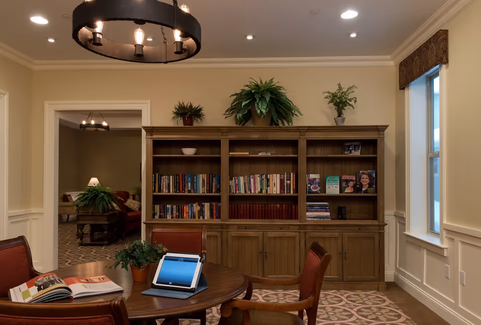 A cozy communal reading room with a large wooden bookshelf, a round table with chairs and a tablet, potted plants, and warm overhead lighting.