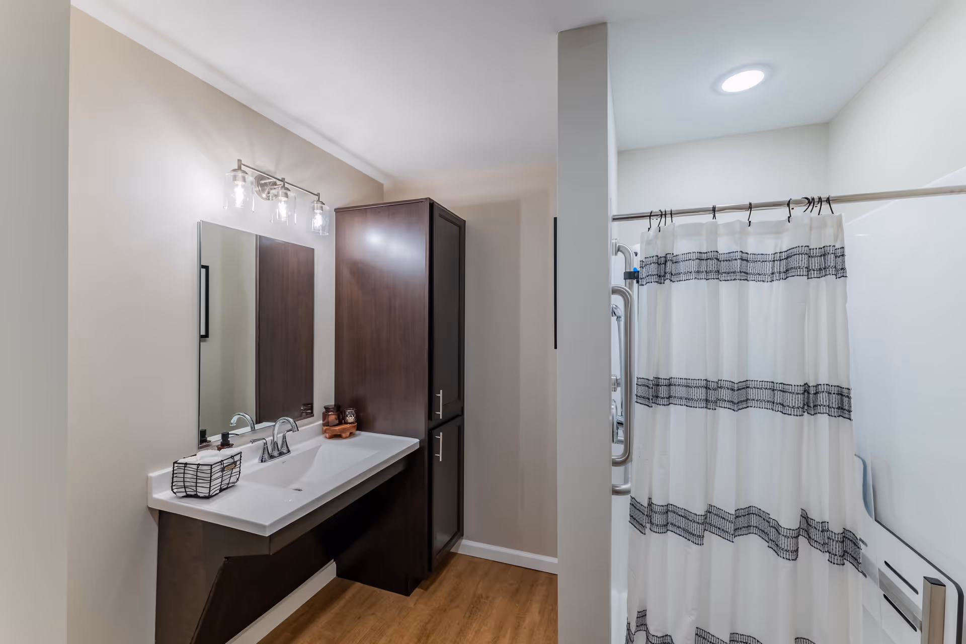 A modern bathroom featuring a white sink with a silver faucet, a large mirror above the sink, and a dark wood cabinet beside it. There is a shower area with a white and gray striped curtain and a grab bar visible. The floor has a wood-like finish and the walls are painted light beige.