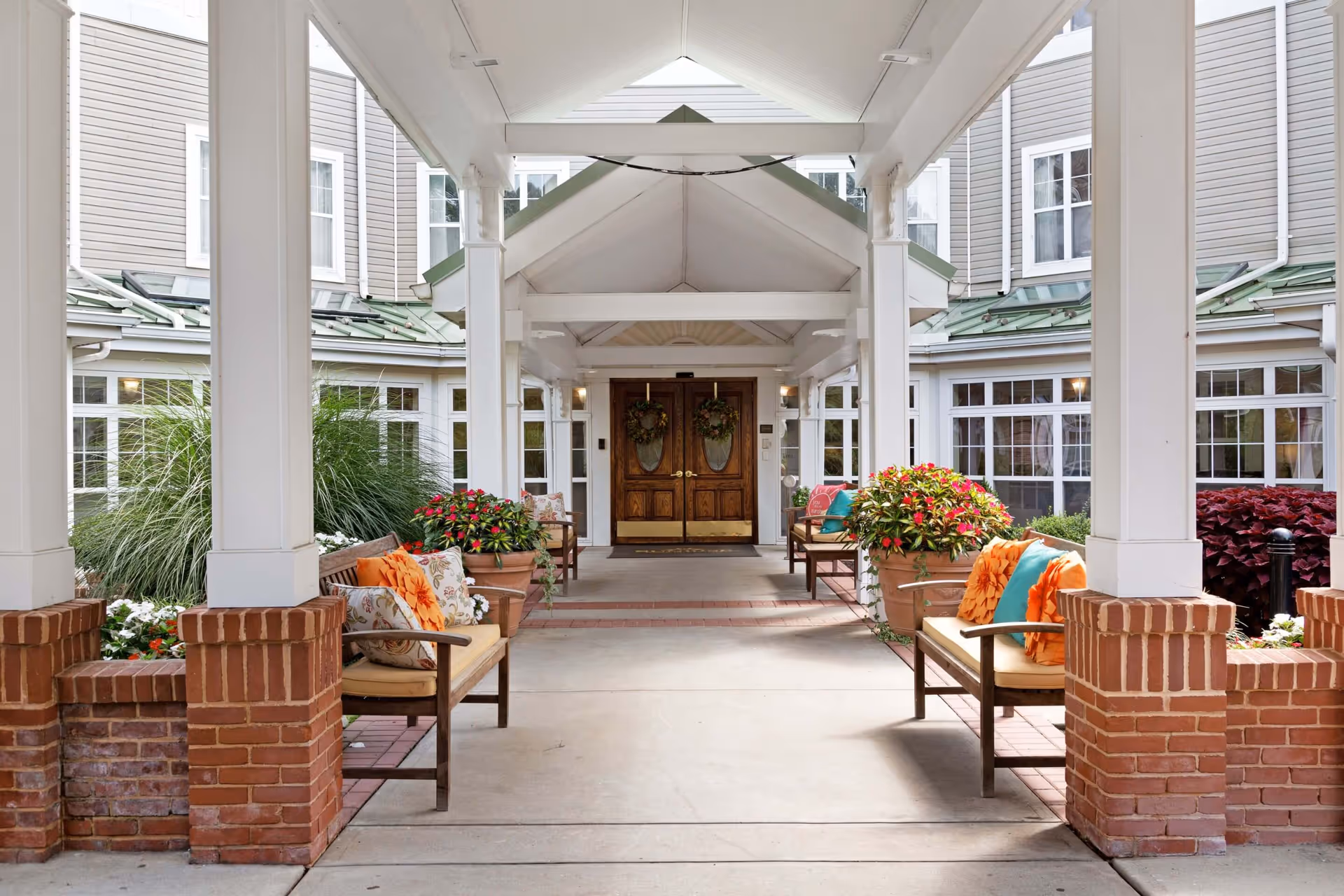 Covered entrance area of a senior living facility with wooden double doors at the end. The walkway is flanked by brick pillars and benches with colorful cushions and potted plants with flowers on both sides. The building has multiple windows and a green roof.
