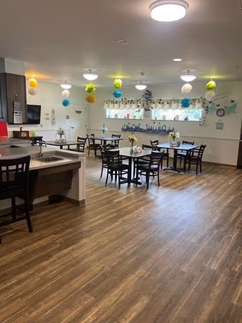 A bright and clean dining area with several tables and chairs arranged on a wood-patterned floor. Colorful hanging decorations are suspended from the ceiling. A kitchen counter with a sink and bar stools is visible on the left side. Windows with floral curtains let in natural light, and the walls are decorated with various small items and artwork.