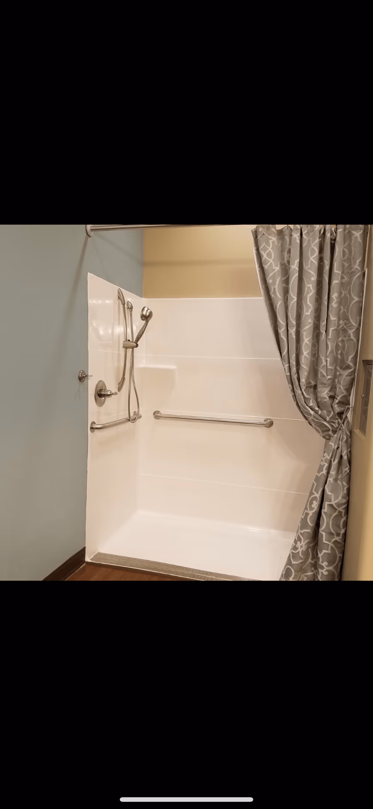 A shower area with a white fiberglass surround, a handheld showerhead attached to a vertical slide bar, and two horizontal grab bars. The shower curtain is patterned in gray and white and is tied back to the right side. The walls around the shower are painted in light blue and beige tones, and the floor outside the shower appears to be wood or wood-like.