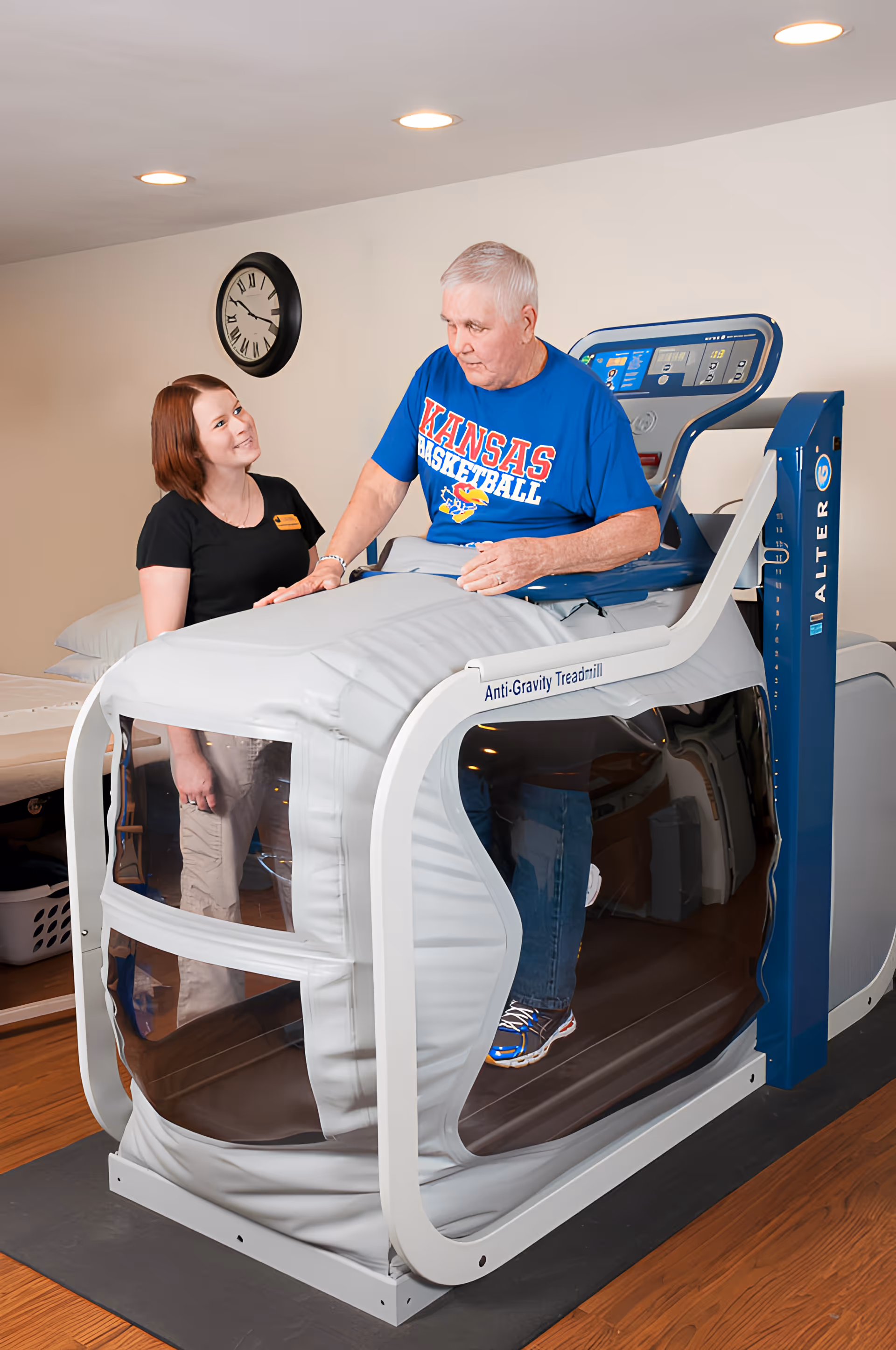 An elderly man wearing a Kansas Basketball t-shirt is using an anti-gravity treadmill while a female staff member stands beside him, smiling. The setting appears to be a rehabilitation or therapy room with a clock on the wall and a treatment table in the background.