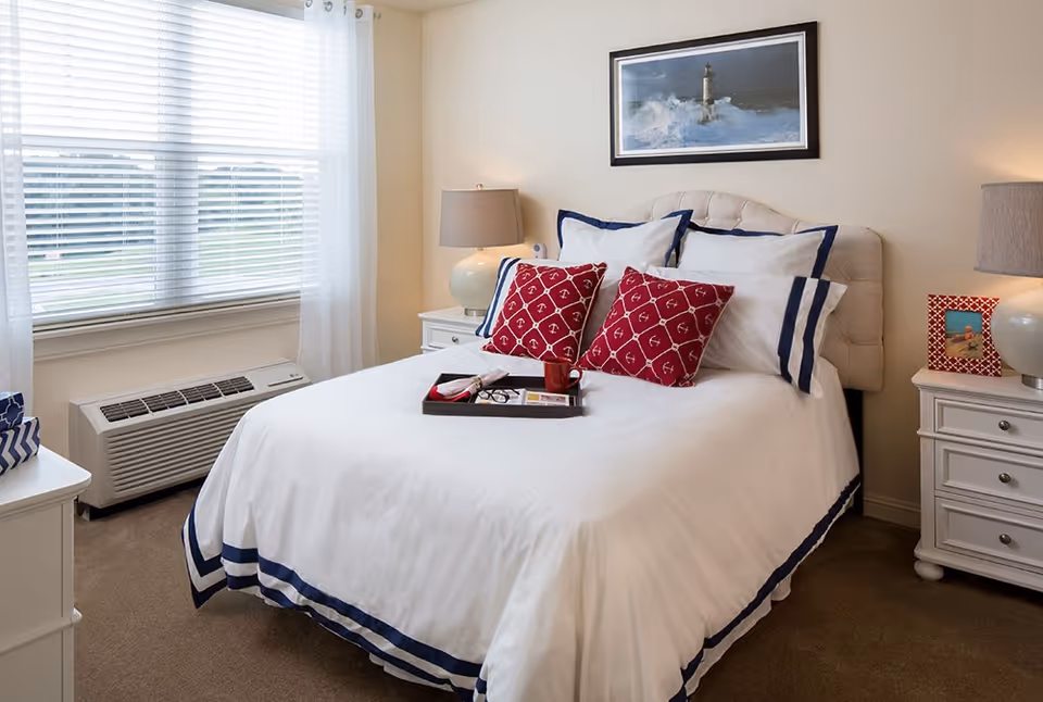 A neatly made bed with white bedding and navy blue trim, adorned with two red decorative pillows with white anchor patterns. The bed has a tufted headboard and is flanked by two white nightstands, each with a lamp. A framed picture of a lighthouse hangs on the wall above the bed. A window with white blinds and sheer curtains is on the left side of the room, and an air conditioning unit is below the window.