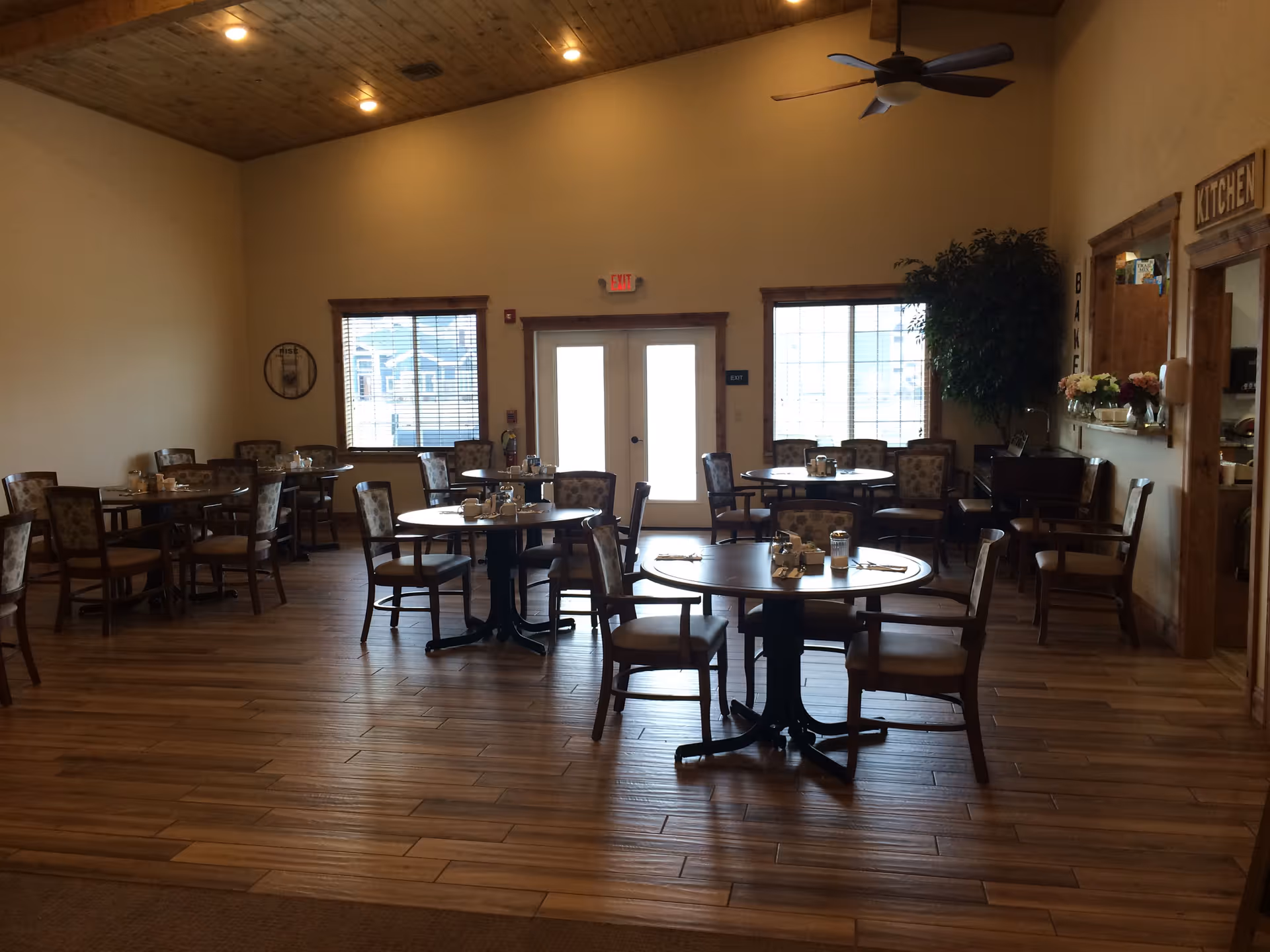 A dining room in an assisted living facility with several round tables and chairs arranged neatly. The room has wooden flooring, a high wooden ceiling with recessed lighting, and large windows letting in natural light. There is a ceiling fan, a door labeled 'Kitchen,' and a large plant in the corner.