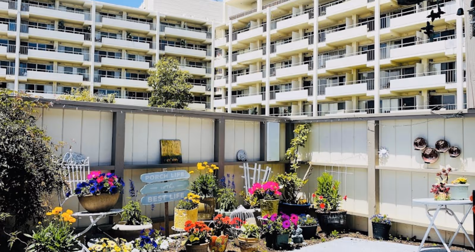 A small outdoor garden area with various colorful potted flowers and plants arranged along a white fence. There is a white metal chair with a flower pot on it, a small white table with plants, and decorative items including a sign that reads 'PORCH LIFE is the BEST LIFE'. In the background, a multi-story building with balconies is visible under a clear sky.
