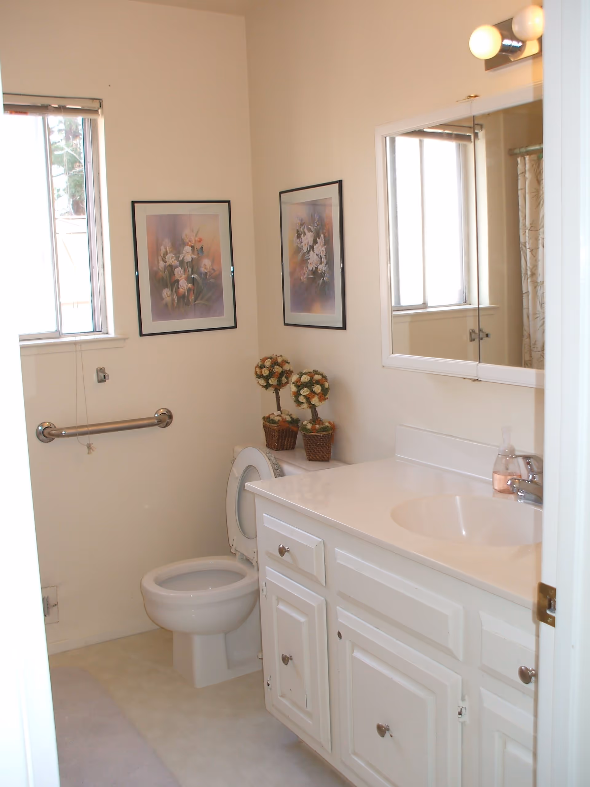 A clean bathroom with a white toilet, a white vanity with a sink, a mirror above the sink, two framed floral pictures on the wall, a window with blinds, a grab bar next to the toilet, and two small potted flower arrangements on the toilet tank.