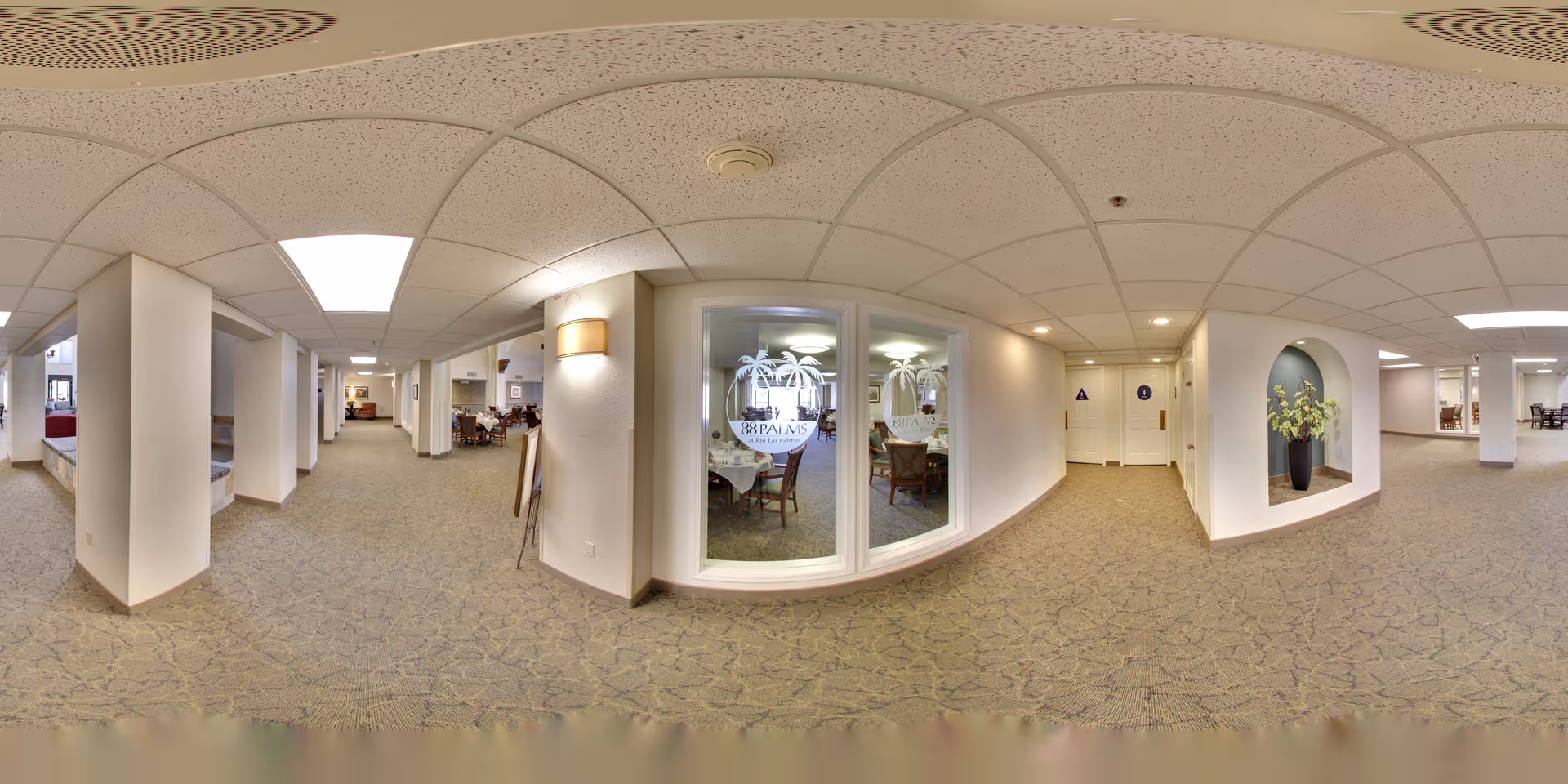 Carpeted interior corridor in a senior living facility with seating areas, decorative alcoves, and a dining room visible through a glass window.