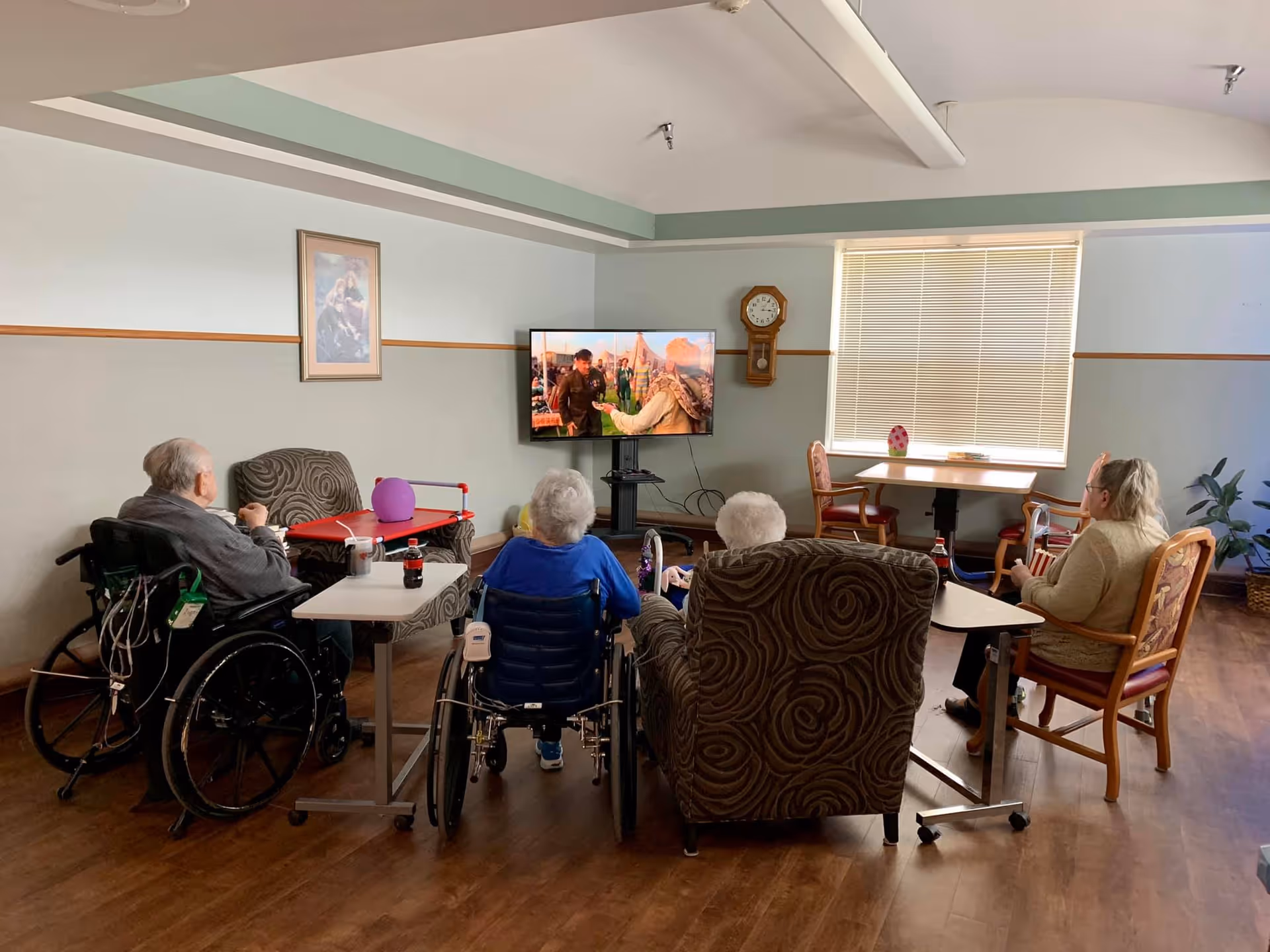 Four elderly individuals, two in wheelchairs and two seated in chairs, watching a television mounted on the wall in a common room with light blue and gray walls, a wooden floor, a clock on the wall, and a window with blinds.