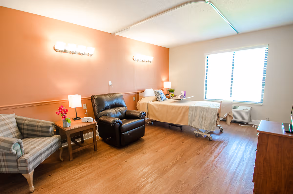 A well-lit senior living bedroom with a hospital-style bed covered in beige bedding, a black recliner chair, a plaid armchair, a wooden side table with a lamp and flowers, and a large window with blinds letting in natural light. The walls are painted in a warm peach and white color, and the floor is wooden.