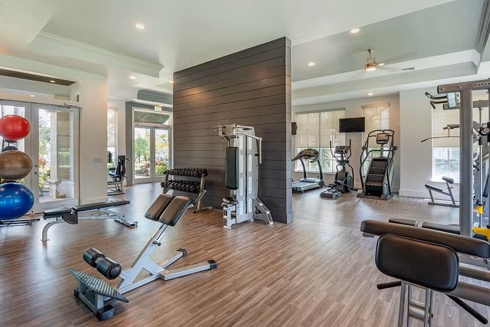Bright, modern fitness room with weight benches, strength machines, cardio equipment, and stability balls on wood-look flooring.