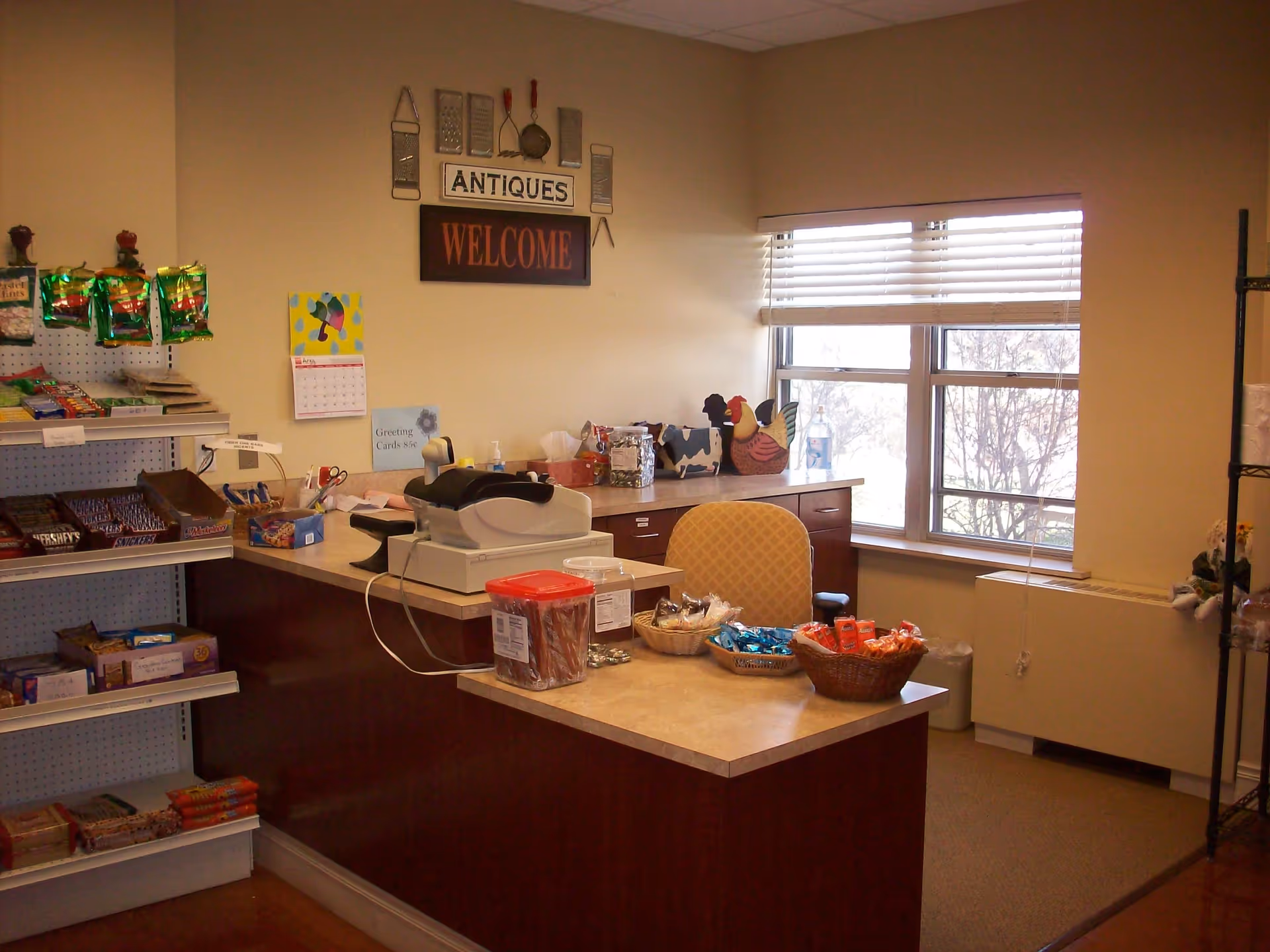 Interior view of a small shop or reception area in a senior living facility with a counter displaying snacks, a cash register, and decorative items. Shelves on the left hold various packaged snacks and candy bars. A window with blinds is visible in the background, along with a yellow patterned chair behind the counter. Wall decorations include kitchen utensils and signs that say 'ANTIQUES' and 'WELCOME'.