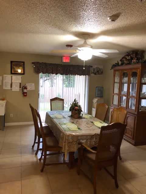 A dining room with a rectangular table covered with a lace tablecloth and six wooden chairs around it. The table has placemats and a floral centerpiece. There is a ceiling fan with a light above the table. In the background, there is a large window with vertical blinds and a valance, a wooden china cabinet with glass doors, a water dispenser, and a fire extinguisher mounted on the wall.