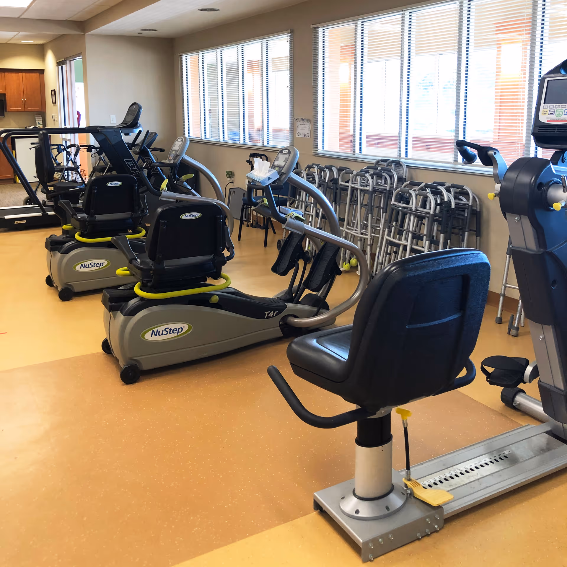 A fitness room with several NuStep exercise machines arranged in rows. The room has large windows with blinds, a beige floor, and a row of walkers lined up against the wall. There is also a black chair attached to one of the exercise machines.