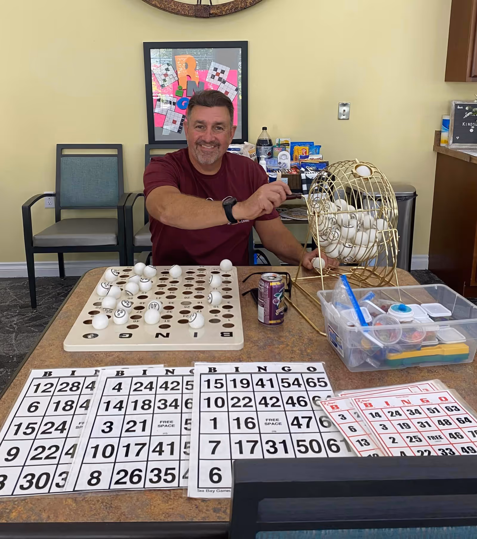 A man sitting at a table playing bingo in a room with yellow walls. The table has bingo cards, a bingo ball cage filled with numbered balls, and a container with various supplies. Behind him are two chairs and a framed colorful artwork on the wall.