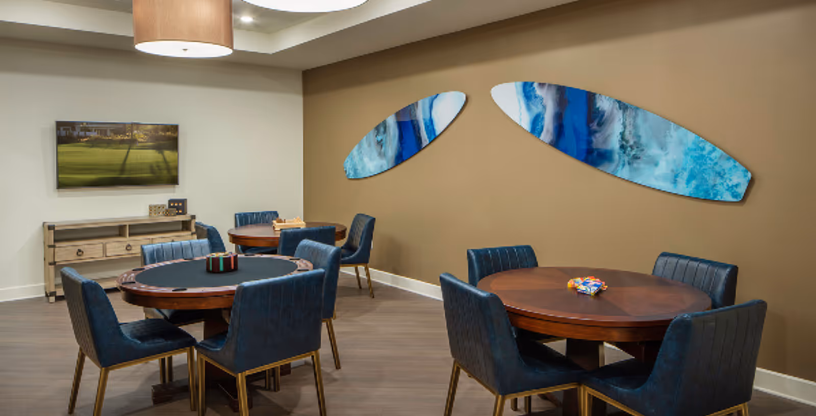 A well-lit interior room with three round wooden tables surrounded by blue upholstered chairs. The tables have board games on them. On the wall, there are two abstract blue and white art pieces. A flat-screen TV is mounted on a white wall above a wooden console table.