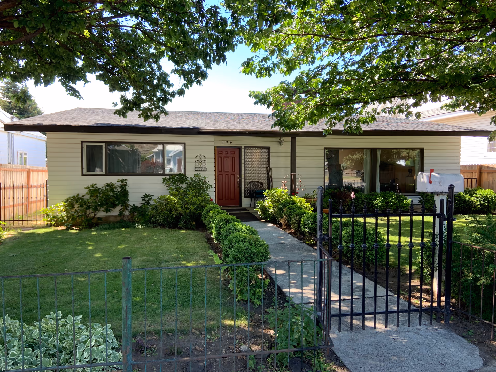 Front exterior view of a single-story house with white siding and a red front door, surrounded by a well-maintained lawn and green bushes. A concrete pathway leads from a black metal gate to the front door. Large tree branches with green leaves hang overhead, casting shade on the house and yard.