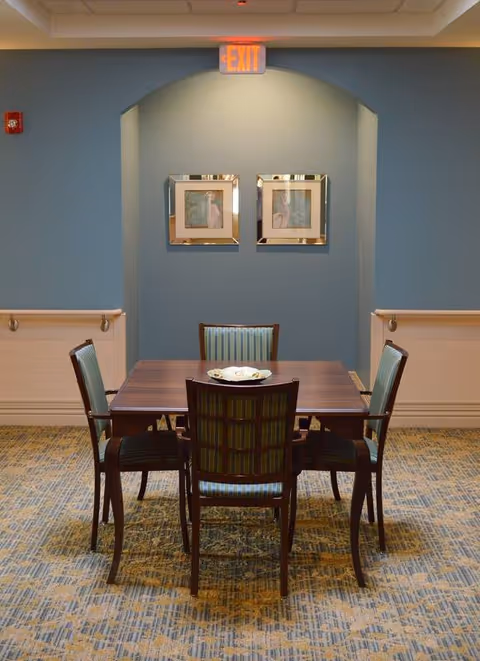 A small dining area with a square wooden table and four striped upholstered chairs. The room has blue walls with a light wood wainscoting and two framed pictures hanging on the wall. An exit sign is illuminated above an arched alcove.