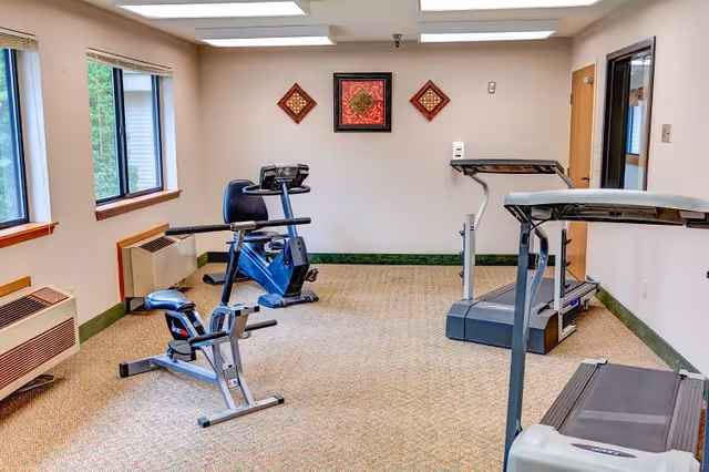 A small exercise room with a treadmill, a stationary bike, and a recumbent bike. The room has beige walls with three decorative wall hangings, carpeted floor, and two windows letting in natural light.