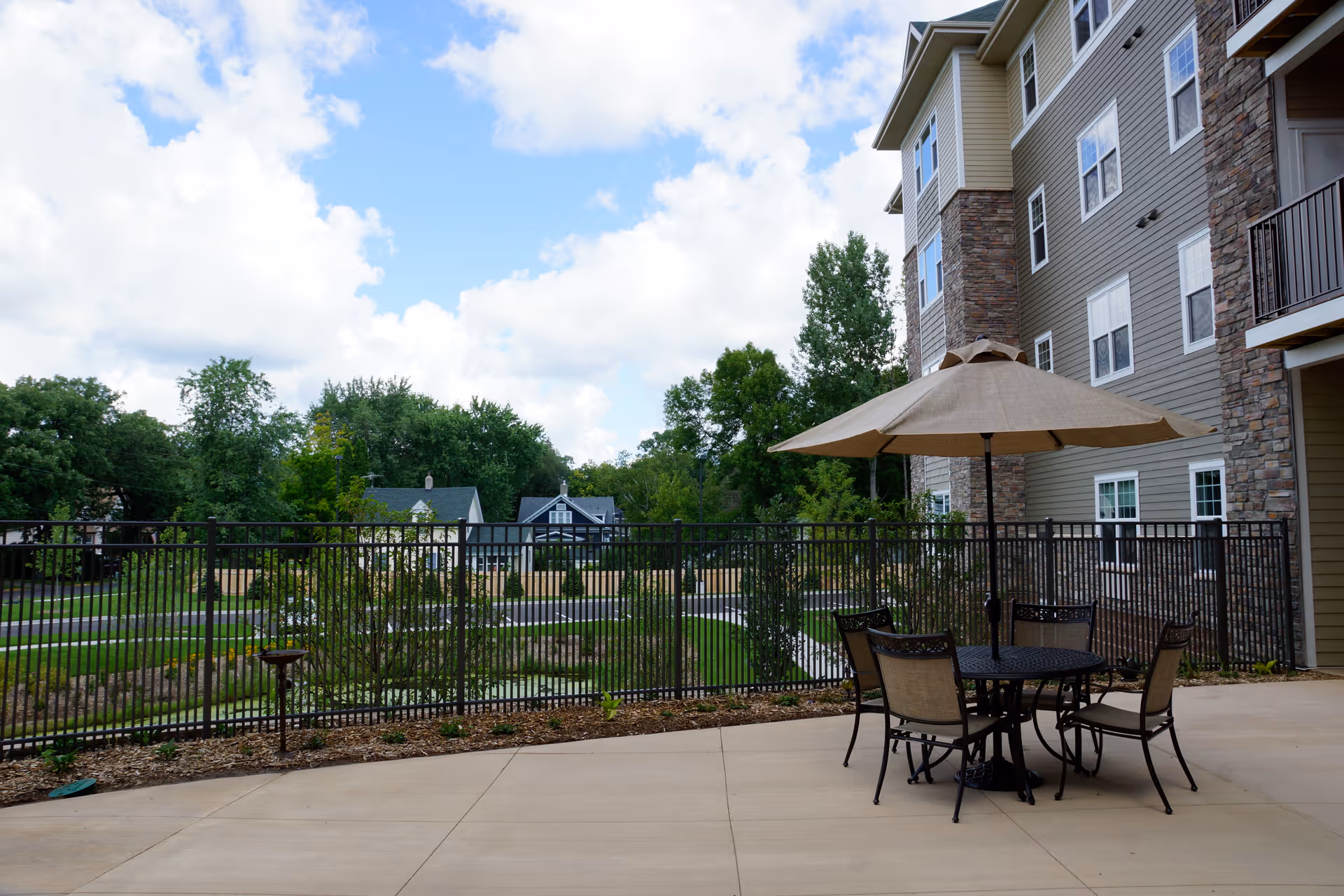 Outdoor patio area at Amira Choice Forest Lake with a round table, four chairs, and a large beige umbrella. The patio is next to a multi-story building with stone and siding exterior. A black metal fence encloses the area, with trees and houses visible in the background under a partly cloudy sky.