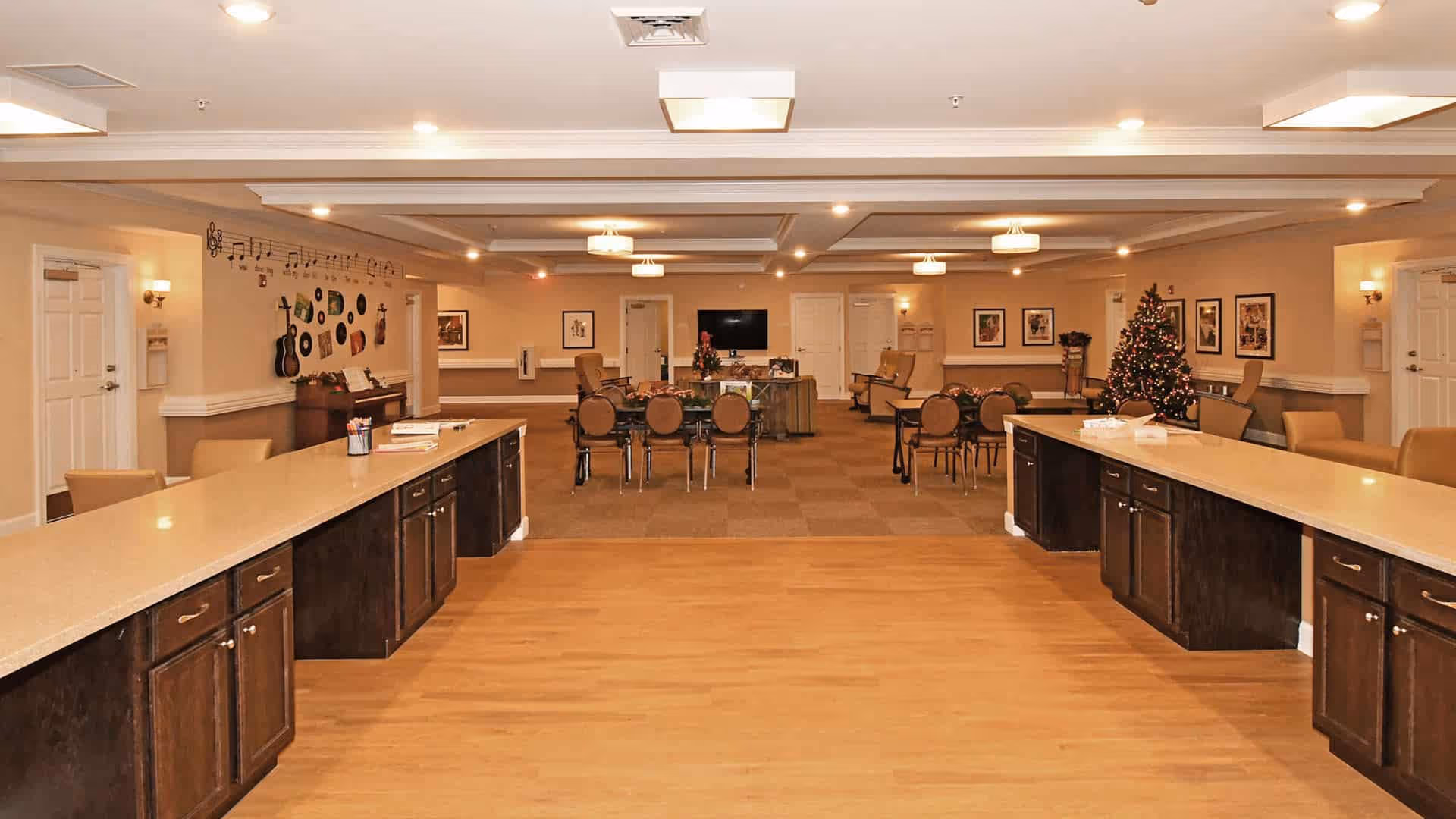 Spacious senior living common/dining room with long service counters in the foreground, tables and chairs and a decorated Christmas tree in the background.