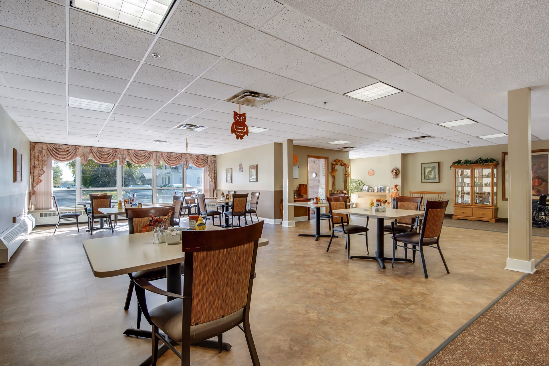 A spacious dining room in an assisted living facility with several tables and chairs arranged for meals. Large windows with decorative curtains allow natural light to fill the room. The room features neutral-colored walls, a tiled floor, and ceiling lights. There are decorative items on the walls and a display cabinet in the background.