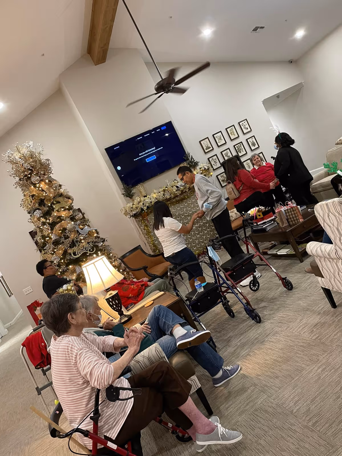 A group of elderly people and caregivers gathered in a decorated living room with a Christmas tree, a large TV mounted on the wall, and framed pictures. Some individuals are seated on chairs and sofas, while others are standing and interacting. The room has a high ceiling with a ceiling fan and warm lighting.