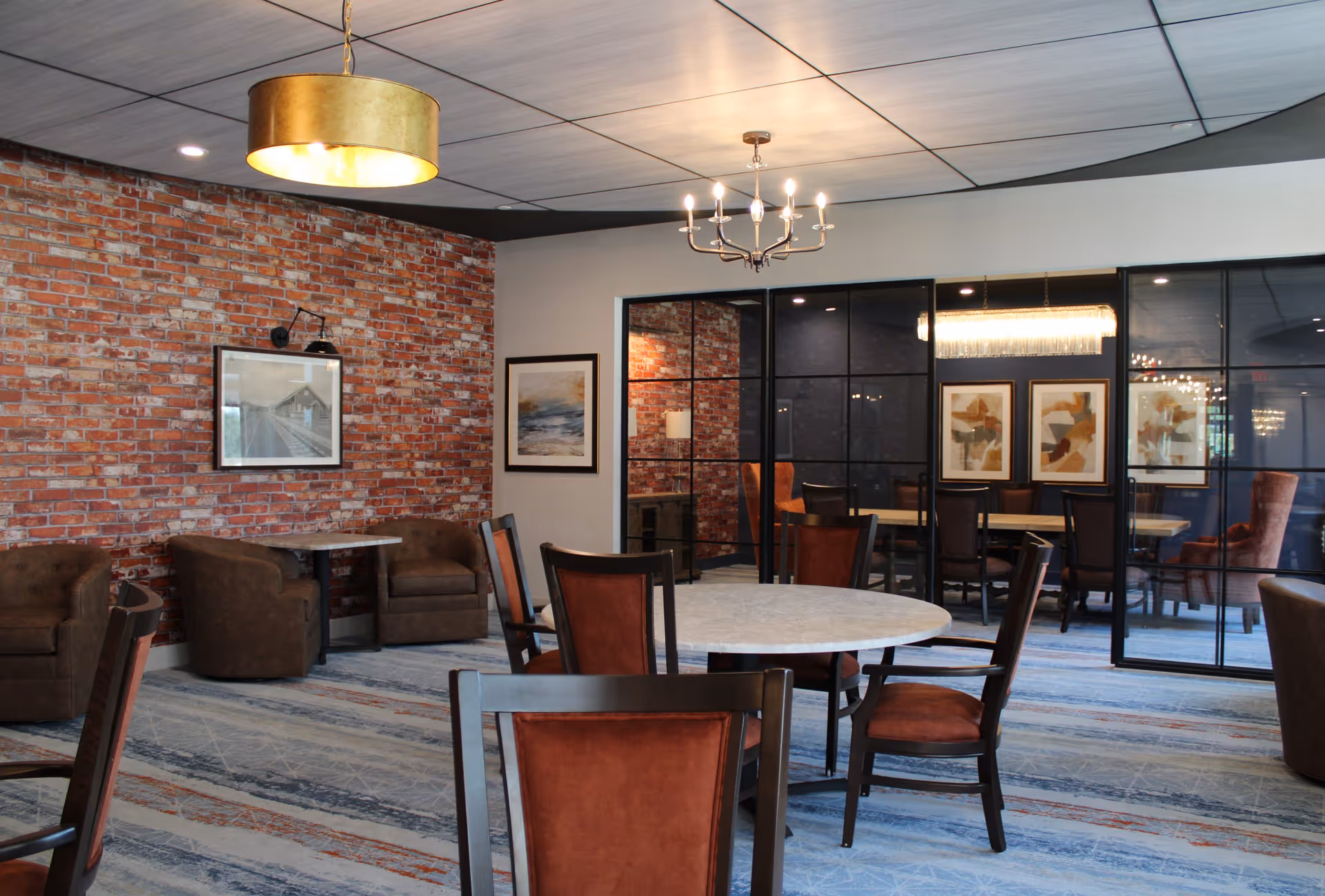 A modern dining room with round tables and chairs featuring brown cushions. The room has a brick accent wall with framed artwork, a blue patterned carpet, and two different ceiling light fixtures including a gold drum pendant and a chandelier. A glass partition separates this area from another room with additional seating and artwork.